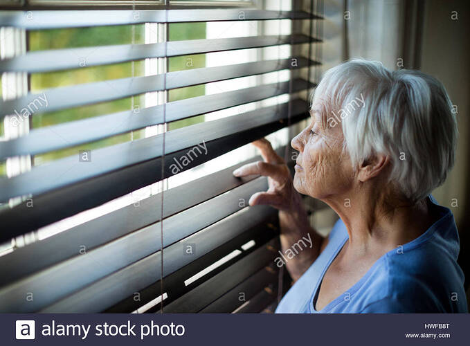 senior-woman-looking-out-from-window-blind-at-home-HWFB8T